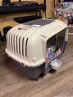 Front and side view of beige and gray hard-sided dog crate with metal door, water bottle, food dish, blanket visible