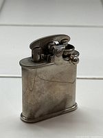 Side view of the antique Colibri lift arm pocket lighter showing the rectangular metal body with visible signs of aging and patina.