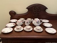 Full set of antique Nippon child's tea service displayed on wooden surface including teapot, cream pitcher, sugar bowl, 6 teacups with saucers, and 6 small plates. Floral hand painted designs with pink and purple flowers.