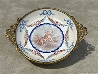 Top-down view of oval dish showing central cherub painting, floral garlands and gilt metal rim and handles.