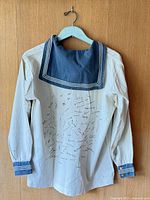 White cotton naval sailors shirt with navy blue square collar and cuffs, hung on wooden background, showing multiple pen signatures on back