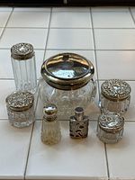 Full view of 7 antique vanity jars arranged on a tiled surface showing crystal and sterling silver lids with designs
