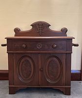 Front view of antique walnut washstand showing two oval burled walnut inset panels on cabinet doors, one drawer with three round wooden knobs, and carved wood top backboard.