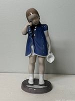Front view of Bing & Grondahl porcelain figurine of a young girl in a blue dress holding a tipped-over white milk jug with visible milk spill on the base.