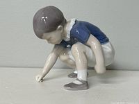 Porcelain figurine of a boy squatting, dressed in blue and white clothing, showing details on the front and side profile.