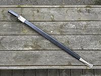 Full length image showing sword sheathed in black leather scabbard with metal tip on wooden outdoor surface.
