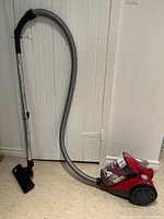 Full view of red Dirt Devil Featherlite canister vacuum with hose extended and floor head attachment upright on linoleum floor near white paneled door.