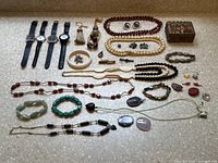 Full view of assorted jewelry including necklaces, bracelets, gemstones, watches, earrings and a small box all laid out on a stone surface