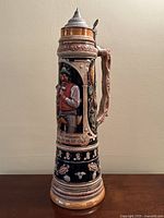 Front view of the tall ceramic beer stein showing the man in traditional attire with detailed colored relief and pewter lid.