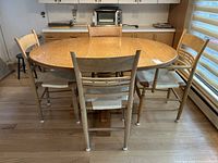 Front and side view of vintage oak breakfast table with four light-colored wooden chairs around it showing the table's oval shape and light wood finish.