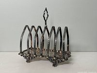 Side view showing ornate feet and lattice base of silver plated toast rack with multiple curved slots for toast slices.