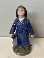 Front view of blue raincoat porcelain figurine holding folded umbrella, standing on grassy base.