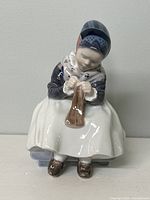 Front view of Royal Copenhagen porcelain figurine of a girl sewing, showing detailed bonnet and dress.