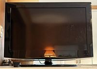 Front view of black Samsung flat screen TV with glossy black bezel and Samsung logo below the screen standing on a black base stand, reflecting light from room.
