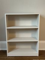 Front view of a small white bookshelf with three shelves placed against a beige wall and white baseboard.