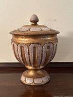Front view of gold and white ceramic Florentine style pedestal jar with lid, showing detailed scalloped and vertical panel design.