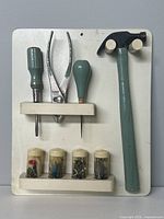 Front view of vintage tool organizer board with four green-handled tools (screwdriver, pliers, awl, hammer) and four small plastic containers with nails and screws.