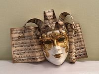 Front view of the mask showing gold detailing on white face and curled sheet music decorations with small bells attached.