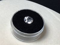 Top-down view of a single clear round faceted Azozeo Phenacite gemstone in a transparent plastic case with black felt interior.
