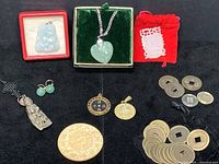 Full lot overview showing green stone heart pendant in box, green stone drop earrings, white carved stone amulet, antique fingernail protector necklace, multiple Asian coins including metal and wooden disks.
