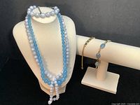 Five-piece jewelry set including baby blue graduated blue lace agate necklace and bracelet on mannequin, two bracelets with gold and black opal links, and blue topaz tennis bracelet on display bar.