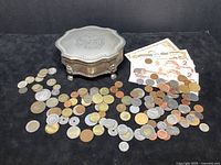 Lot overview showing metal Victorian style jewelry music box surrounded by assorted foreign coins and paper bills.