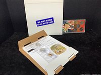 Photo showing the white mailer envelope marked 'DO NOT BEND OPEN WITH CARE' with the Kennedy Centennial Collection coins, stamps, and ephemera set inside, along with a display card showing John F. Kennedy and the coins.