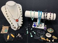 Wide view of assorted mineral and stone pendants, necklaces, bracelets displayed on necklace stands and flat black surface. Includes turquoise bracelets, glass jar pendant, chakra necklace, and other assorted mineral jewelry.