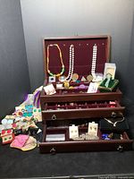 Full view of open three-tier wooden jewelry box with numerous jewelry pieces including necklaces, bracelets, brooches, earrings on and inside the box.
