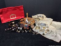 Wide shot showing all jewelry pieces including red satin Christian Dior pouch, wooden ring holder, watches, scarves, necklaces, earrings, brooches, pendants and bracelets arranged on black background.