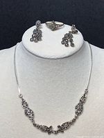 Front view of three sterling silver and marcasite jewelry pieces displayed on a black and white stand: necklace, ring, and dangle earrings.