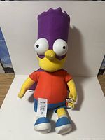 Full view of large plush Bart Simpson toy sitting upright, showing character details including oversized eyes, red shirt, blue shorts, and shoes.