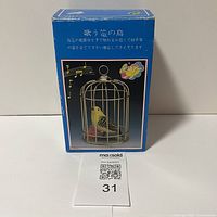 Front view of the original box showing the yellow bird in a metal cage swinging. Box is blue with Japanese text and a musical note icon, indicating the bird may produce sounds.