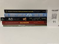Stack of various books including topics like popular music culture, Queen biography, The Script book, and Buffy the Vampire Slayer script book.