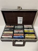 Top-down photo of open black carrying case holding 24 assorted cassette tapes arranged in rows.