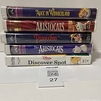 Stacked spines of five Disney VHS movies showing titles: Alice in Wonderland, The Aristocats (two copies), Pinocchio, and Discover Spot with visible case wear.