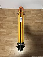 Full view of the collapsed surveying tripod showing three long yellow and orange legs with sharp metal tips and a carrying handle.
