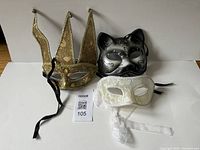 Three masquerade masks displayed on a white surface: gold jester, silver cat, white lace.