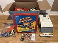 Box with opened flaps showing a red Micro Minis Collector's Case, various small vehicles in plastic bags, and a large white Micro Machines bus toy with rainbow stripes.