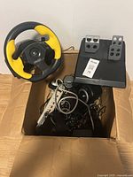 Logitech racing wheel with yellow grips and pedals shown inside the cardboard box along with wires and a power strip.