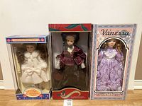 Three new in box porcelain dolls with detailed period costumes in different colored dresses and hats.