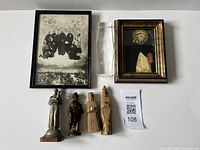 Photo showing framed religious prints, two metal statues, two wooden carvings, and a clear glass vase.