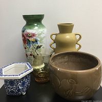 Five vases shown together including the tall floral hand painted antique Bristol glass vase in green, yellow geometric ceramic vase with handles, amber art glass vase, brown planter vase with floral embossing, and blue and white ceramic plant pot with three legs.
