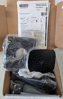Opened box showing the matte black shower head and components in plastic packaging with instruction manual.