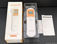 Photo showing the white and orange Landwind LW FT116 digital infrared thermometer, box with image and model name, thermometer in clear plastic holder and instruction sheet beside it.
