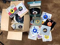 Box of assorted 45 RPM vinyl records, many in paper sleeves, some loose, and a small green carrying case holding some records.
