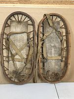 Top-down view of pair of wooden snowshoes on plain surface showing wood frame, wooden crossbars, fabric base, and foot straps.