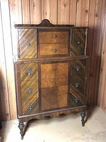 Front view of the antique tallboy dresser showing upper small doors, drawers, and lower larger drawers with detailed wood veneer pattern.