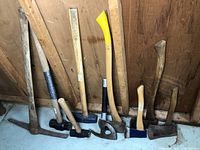 Eight tools arranged vertically against wooden wall: pick/mattock, small pick hammer, two blue sledgehammers, splitting maul, framing axe and hatchet