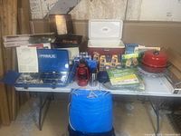 Photo of camping lot contents on a folding table showing Primus stove, coolers, tarps, lantern, convection cooker, and various camping supplies.
